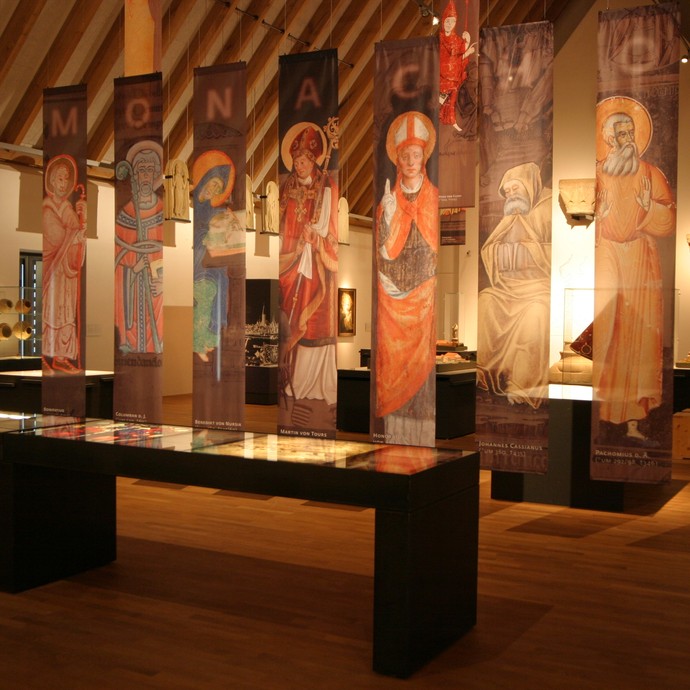 Ein Ausstellungsraum mit Vitrinen im Hintergrund. Von der Decke hängen längliche Fahnen, auf denen damalige Mitglieder des Klosters abgebildet sind. (vergrößerte Bildansicht wird geöffnet)