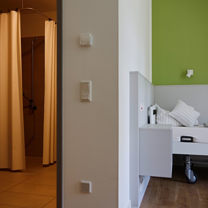 Links gibt eine geöffnete Tür den Blick auf ein Badezimmer frei, rechts steht ein Bett vor einer grünen Wand. (vergrößerte Bildansicht wird geöffnet)