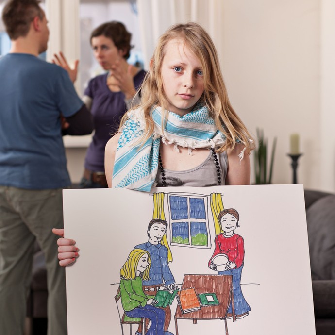 Ein Mädchen schaut traurig, während sich ihre Eltern hinter ihr streiten. Sie hält ein Schild hoch, dass eine glückliche Familie zeigt. (vergrößerte Bildansicht wird geöffnet)